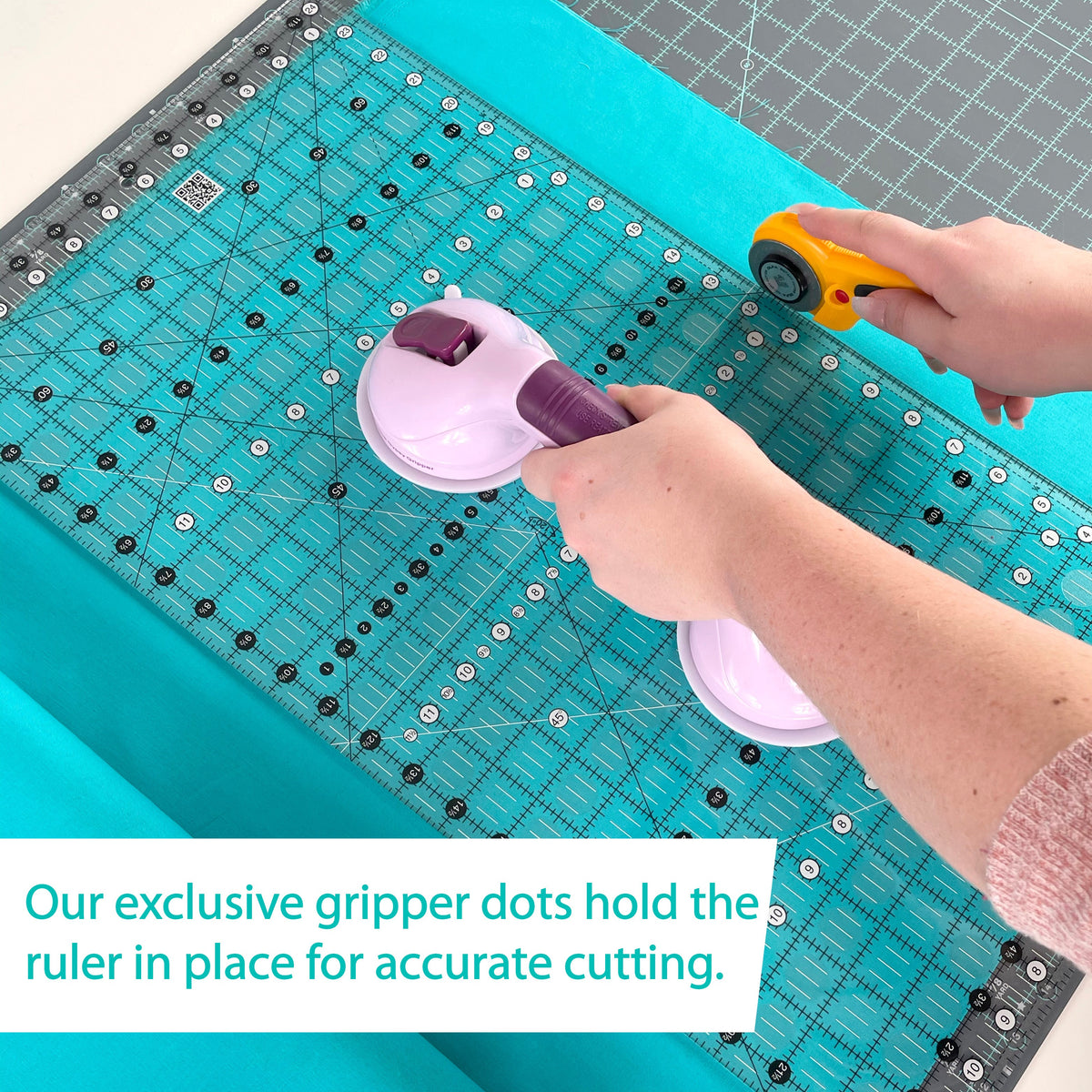 Hands cutting teal fabric along the Creative Grids 12-1/2 x 24-1/2 inch ruler CGR1224 with a suction-cup ruler handle, with a brand caption about the gripper dots holding the ruler in place.