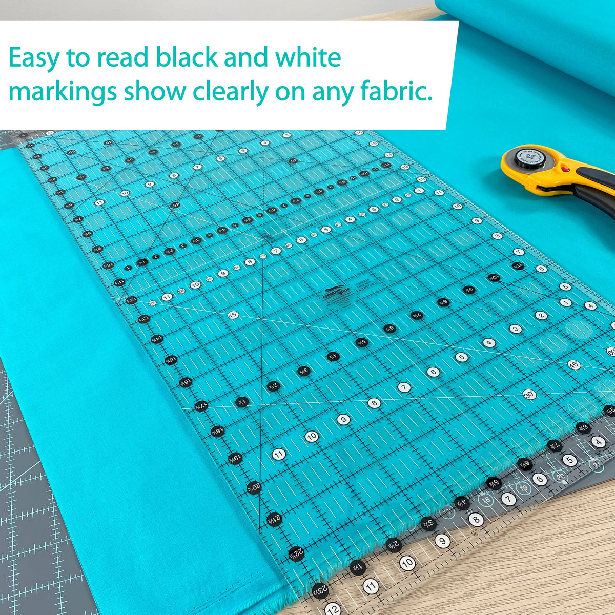 Creative Grids 12-1/2 x 24-1/2 inch ruler CGR1224 laid on teal fabric beside a rotary cutter, with a brand caption about the black and white markings showing on any fabric.