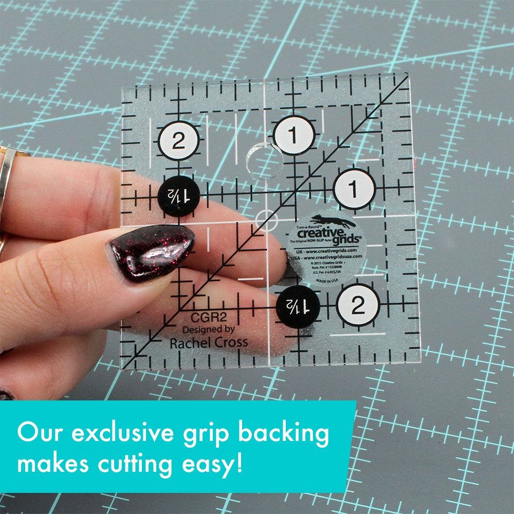 Creative Grids 2-1/2 inch square ruler CGR2 held between finger and thumb over a gray cutting mat, with the frosted non-slip grip visible through the clear acrylic and a caption about the exclusive grip backing.