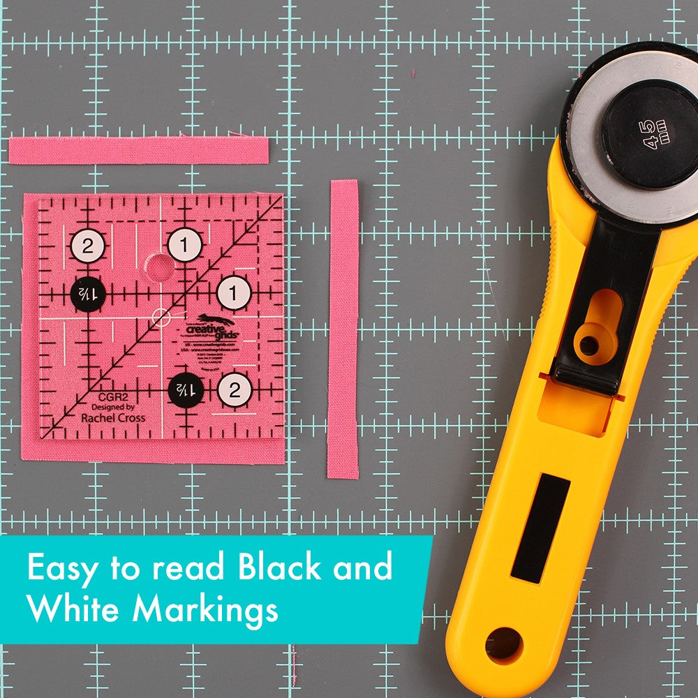 Creative Grids 2-1/2 inch square ruler CGR2 on pink fabric on a cutting mat next to a rotary cutter, captioned Easy to read Black and White Markings.