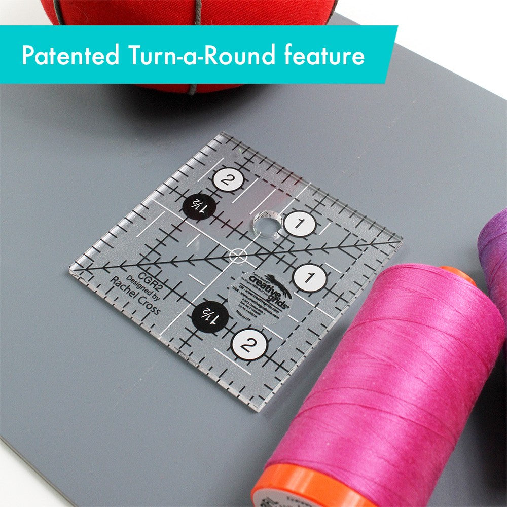 Creative Grids 2-1/2 inch square ruler CGR2 turned on a gray mat beside a spool of pink thread, captioned Patented Turn-a-Round feature.