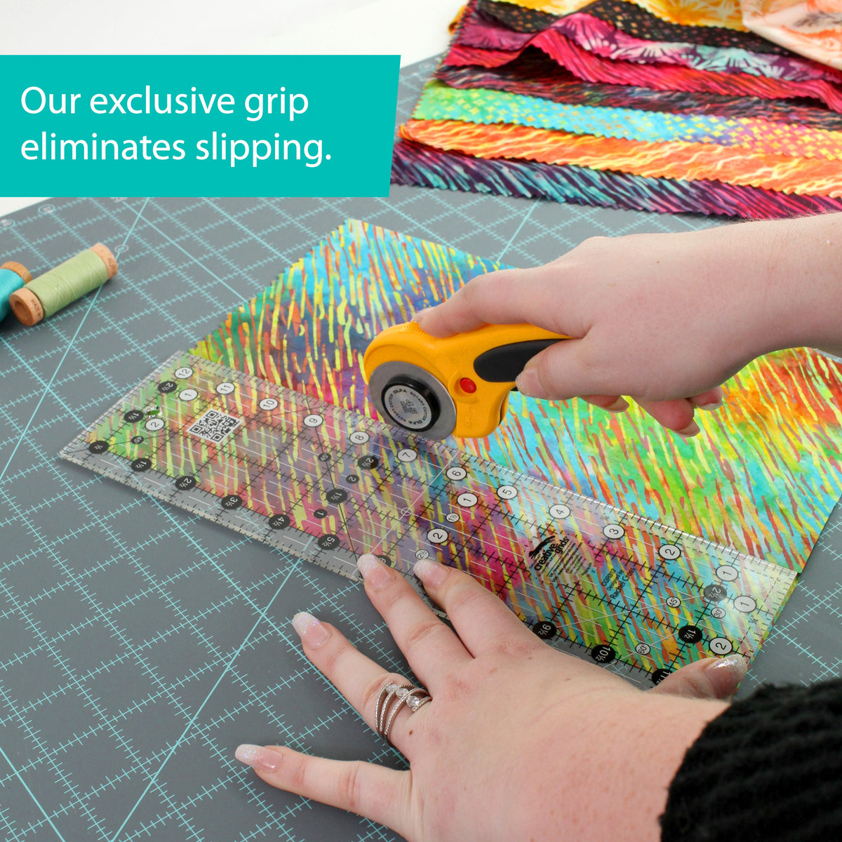 Hands pressing the Creative Grids 3-1/2 x 12-1/2 inch ruler CGR312 on batik fabric while a rotary cutter trims the edge, with a brand caption about the grip eliminating slipping.