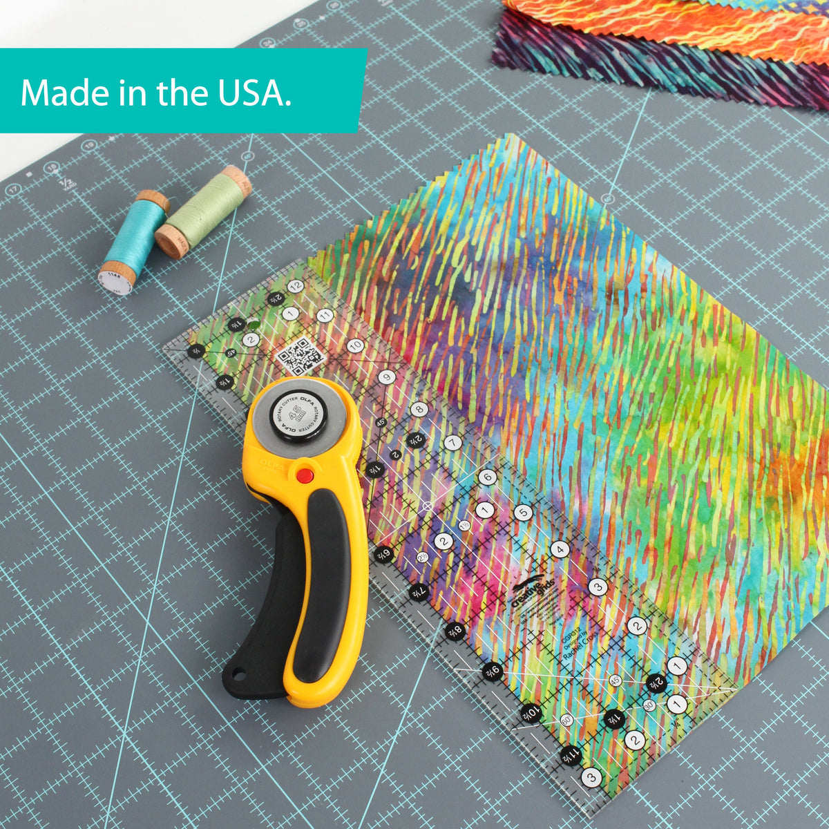 Creative Grids 3-1/2 x 12-1/2 inch ruler CGR312 on batik fabric beside a rotary cutter and spools of thread, with a brand caption reading Made in the USA.