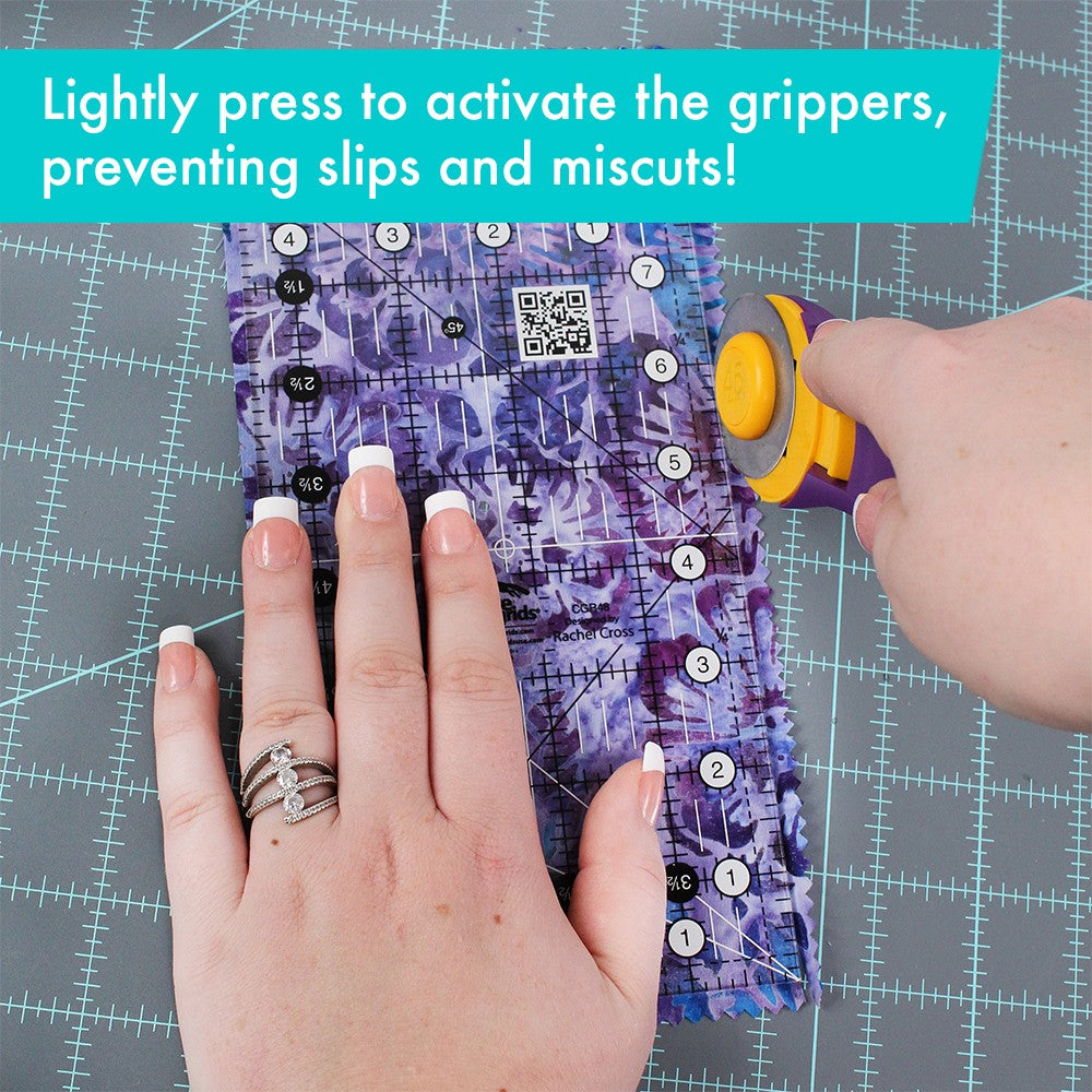 Hand pressing the Creative Grids 4-1/2 x 8-1/2 inch ruler CGR48 on purple batik fabric while a rotary cutter trims beside it, with a brand caption about pressing lightly to activate the grippers.