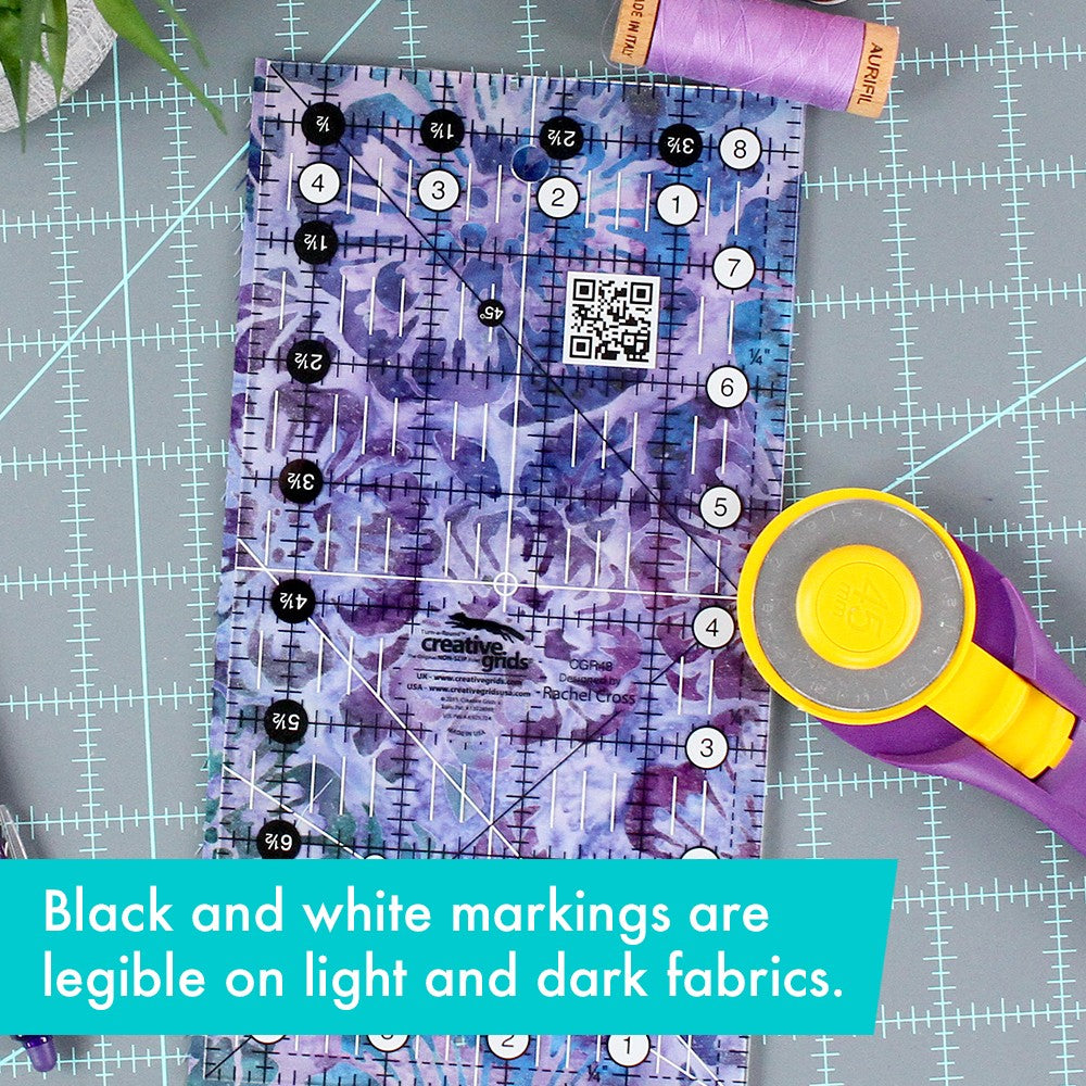 Creative Grids 4-1/2 x 8-1/2 inch ruler CGR48 on purple batik fabric beside a rotary cutter, with a brand caption about the markings reading on light and dark fabrics.