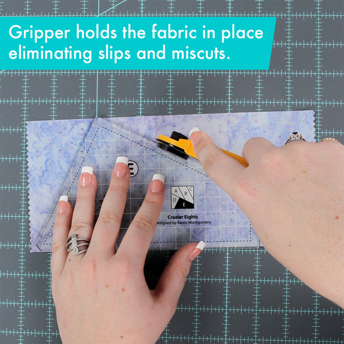 A person holds template E of the Creative Grids Crazier Eights Templates CGRMT7 on a piece of pale blue batik fabric with one hand and cuts along its angled edge with a rotary cutter in the other, on a gray cutting mat. A caption says the gripper holds the fabric in place.