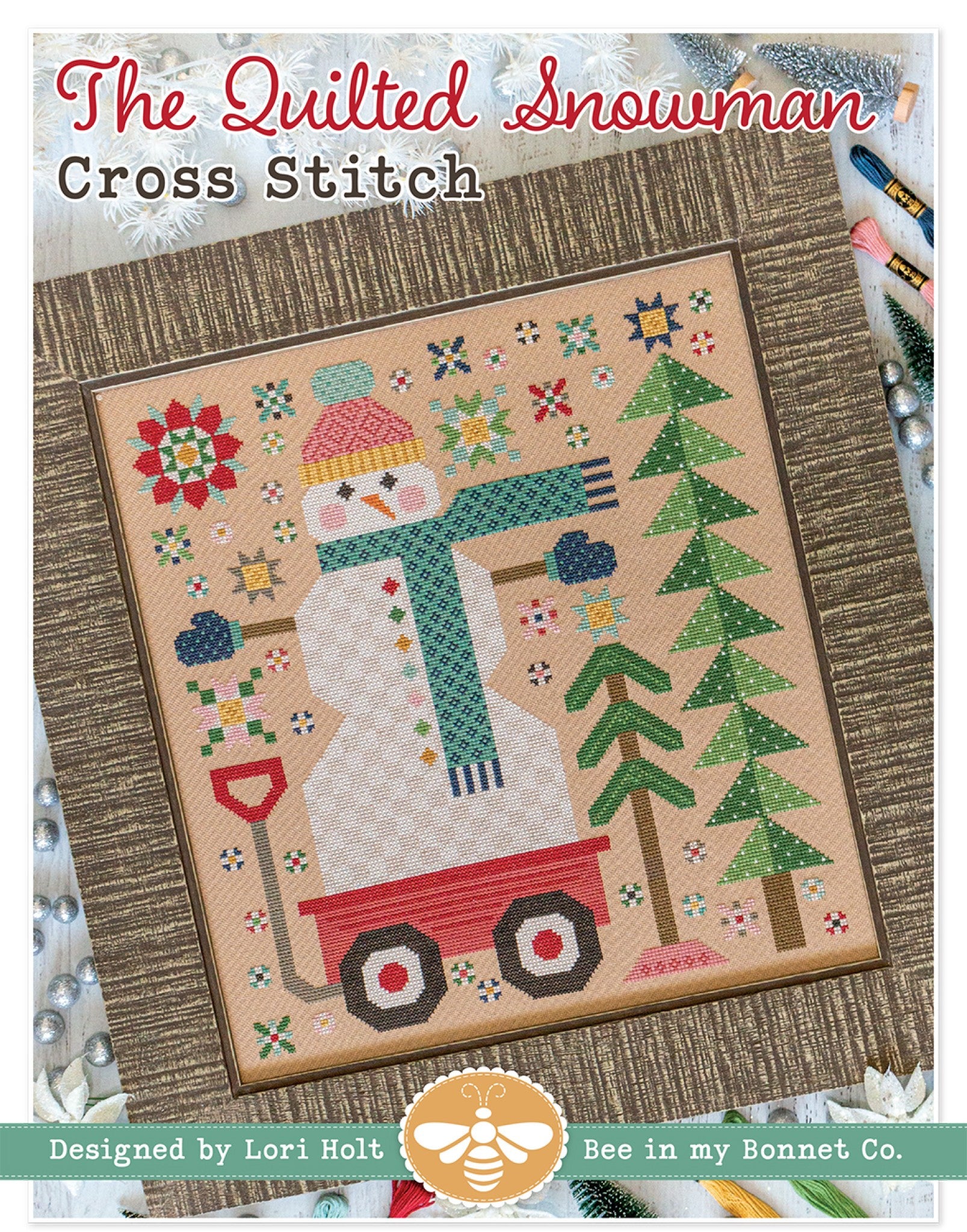 Front cover of The Quilted Snowman cross stitch pattern by Lori Holt for It's Sew Emma. A framed cross stitch shows a snowman in a pink-and-yellow pom-pom hat, teal scarf and blue mittens riding in a red wagon beside green pine trees, with quilt-block stars, snowflakes and a red snow shovel on a pebble-tan background.