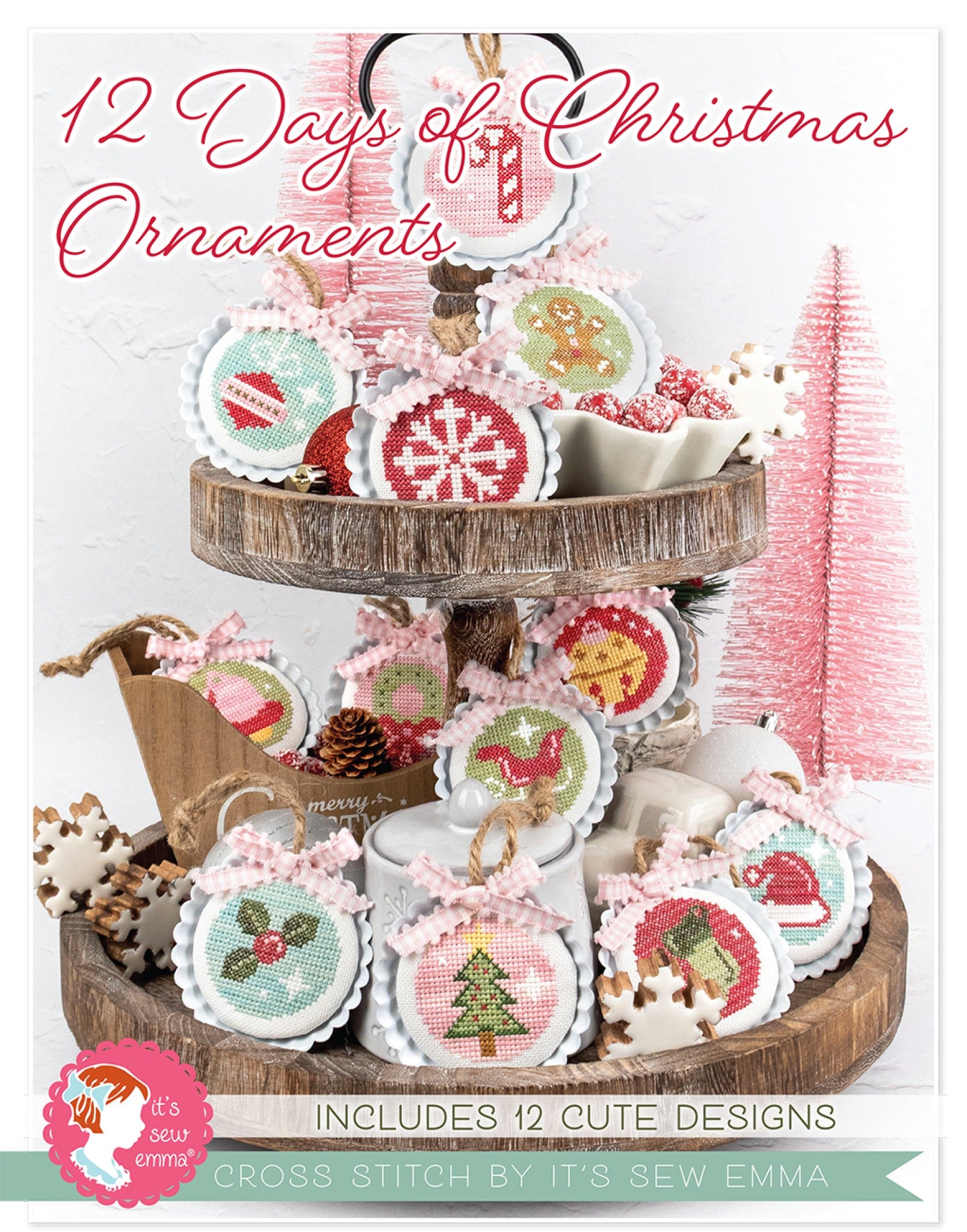 Front cover of the 12 Days of Christmas Ornaments cross stitch pattern by Lori Holt for It's Sew Emma. A two-tier wooden stand displays a dozen round cross stitch ornaments tied with pink gingham bows and twine, including a candy cane, snowflake, gingerbread man, Christmas tree, striped ornament, holly, a red bird, a bell and a Santa hat, worked in soft red, pink, green and blue on cream fabric and styled with pink bottle-brush trees. Text reads Includes 12 Cute Designs.