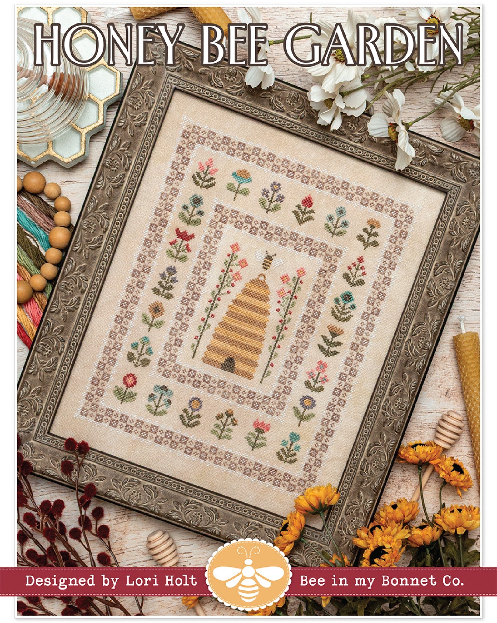 Front cover of Honey Bee Garden cross stitch pattern by Lori Holt for It's Sew Emma. Framed stitched sampler with a tall straw beehive at the center, a bee above the entrance, and tall stems of red and pink flowers on each side. Rows of small folk-art flowers and two nested checkerboard borders with lace-like edges surround the central scene, worked in a muted vintage palette on cream linen. Styled with daisies, beeswax candles, and a honey dipper.
