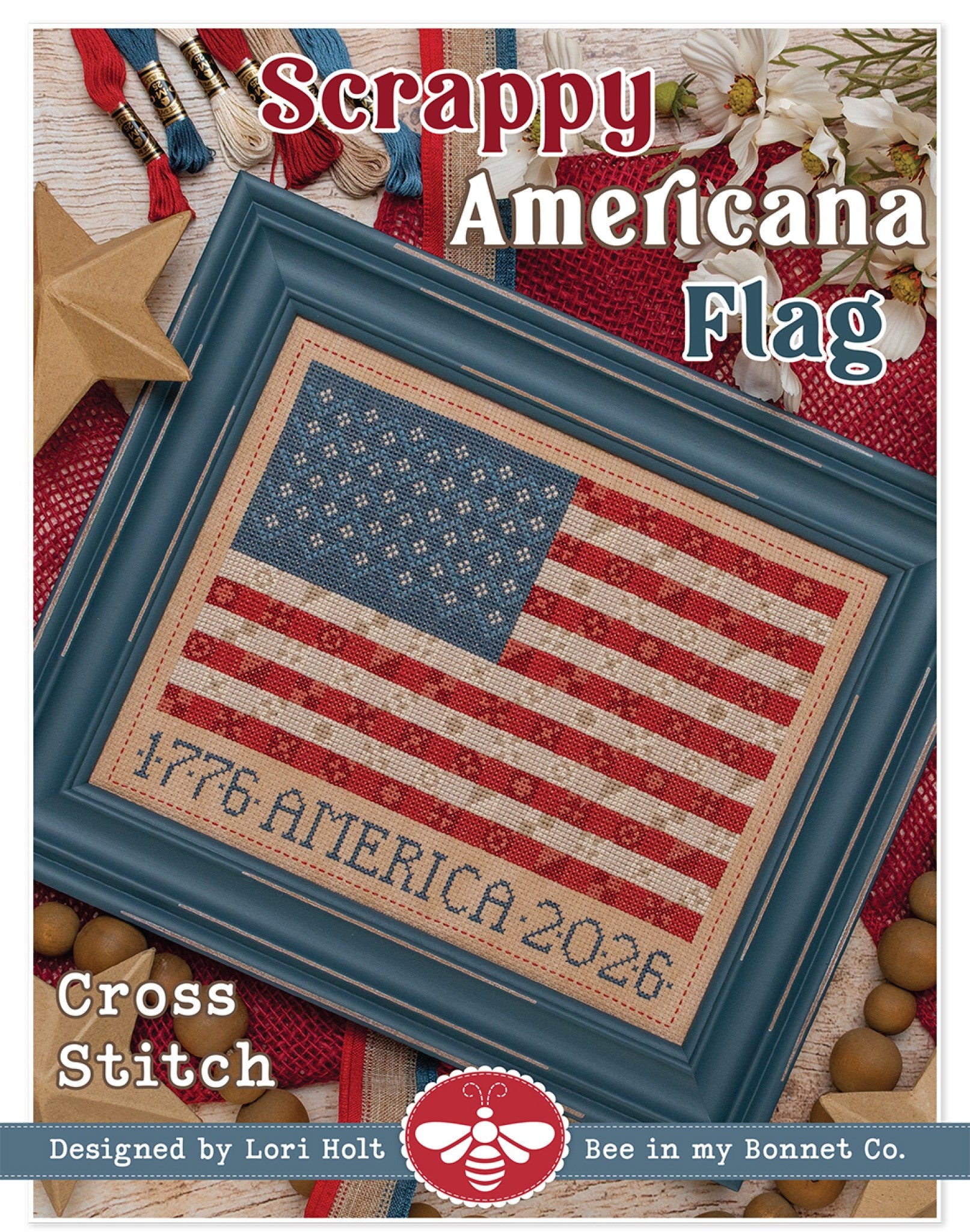 Front cover of Scrappy Americana Flag cross stitch pattern by Lori Holt for It's Sew Emma. A framed cross stitch of the US flag with stars and stripes built from small mottled motifs in faded red, cream and denim blue, with stitched lettering reading 1776 AMERICA 2026, displayed in a distressed blue-gray frame and styled with floss and white flowers.