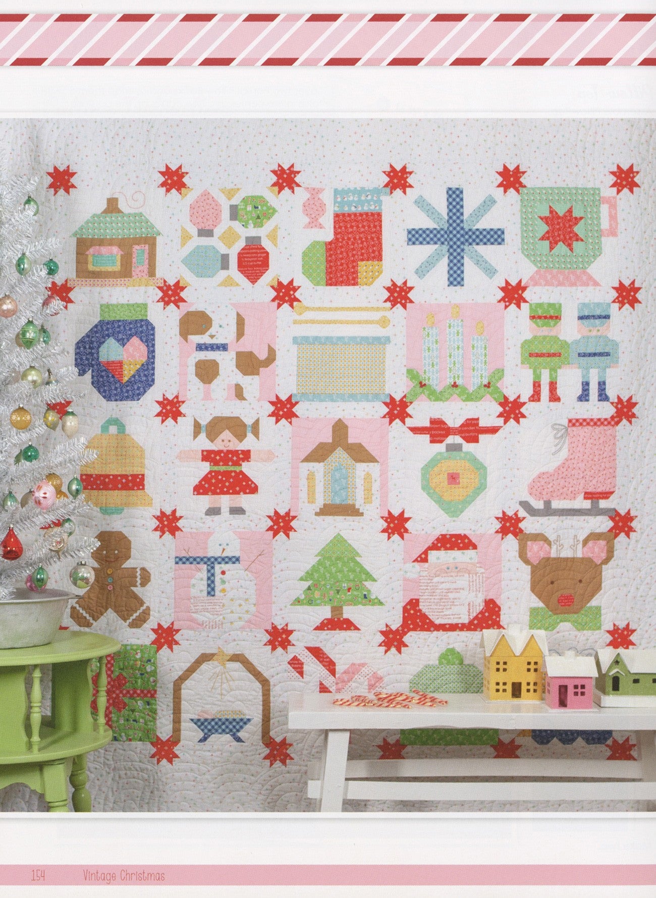 A pink-background version of the Vintage Christmas Sampler from Vintage Christmas by Lori Holt for It's Sew Emma, with red sashing stars and holiday picture blocks, styled beside a white feather tree with vintage ornaments and small glitter houses.