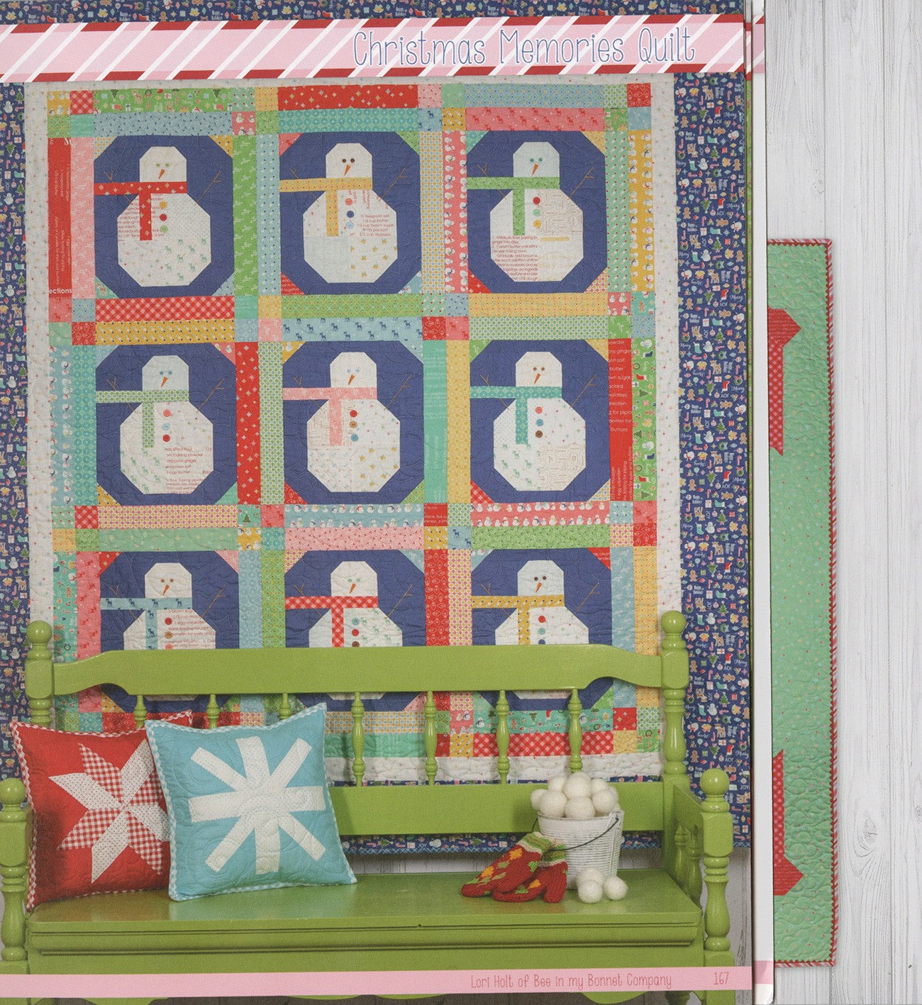 Christmas Memories Quilt from Vintage Christmas by Lori Holt for It's Sew Emma, nine pieced snowman blocks on navy backgrounds with scrappy sashing, draped over a green bench with a red star pillow and an aqua snowflake pillow.