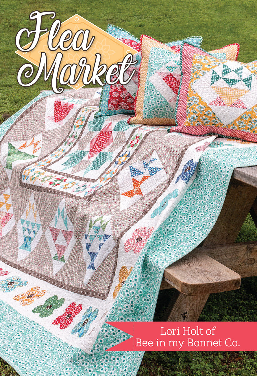The Flea Market medallion quilt by Lori Holt for It's Sew Emma draped over a wooden bench outdoors with two coordinating pillows, showing the scrappy basket and flower blocks around the central star.