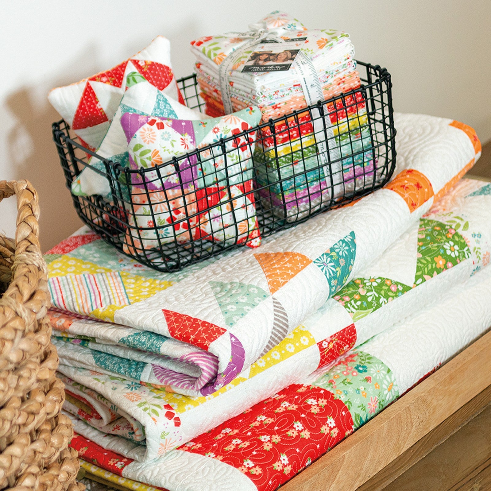 Folded Family Favorites quilts stacked beside a wire basket of coordinating pillows and pincushions.