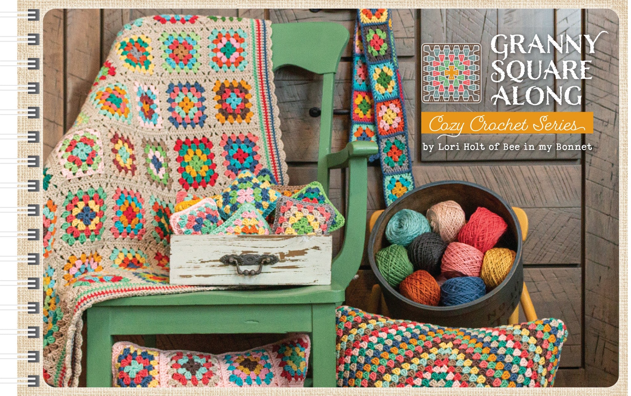 Granny Square Along Crochet Pattern Book cover by Lori Holt of Bee in my Bonnet for It's Sew Emma. A multicolor granny square afghan drapes over a green vintage chair beside a hanging granny square scarf, a black bowl filled with balls of colorful cotton thread, a distressed white box of folded granny squares, and granny square pillows. The title and Cozy Crochet Series logo appear at upper right.