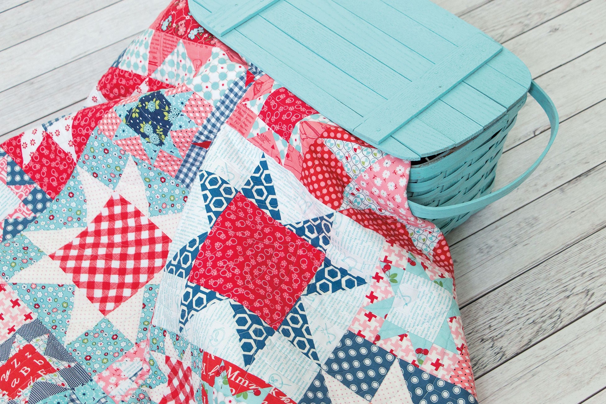 A red, white, and blue scrappy patchwork star quilt from Farm Girl Vintage by Lori Holt for It's Sew Emma, with a vintage teal picnic basket on a whitewashed wood floor.