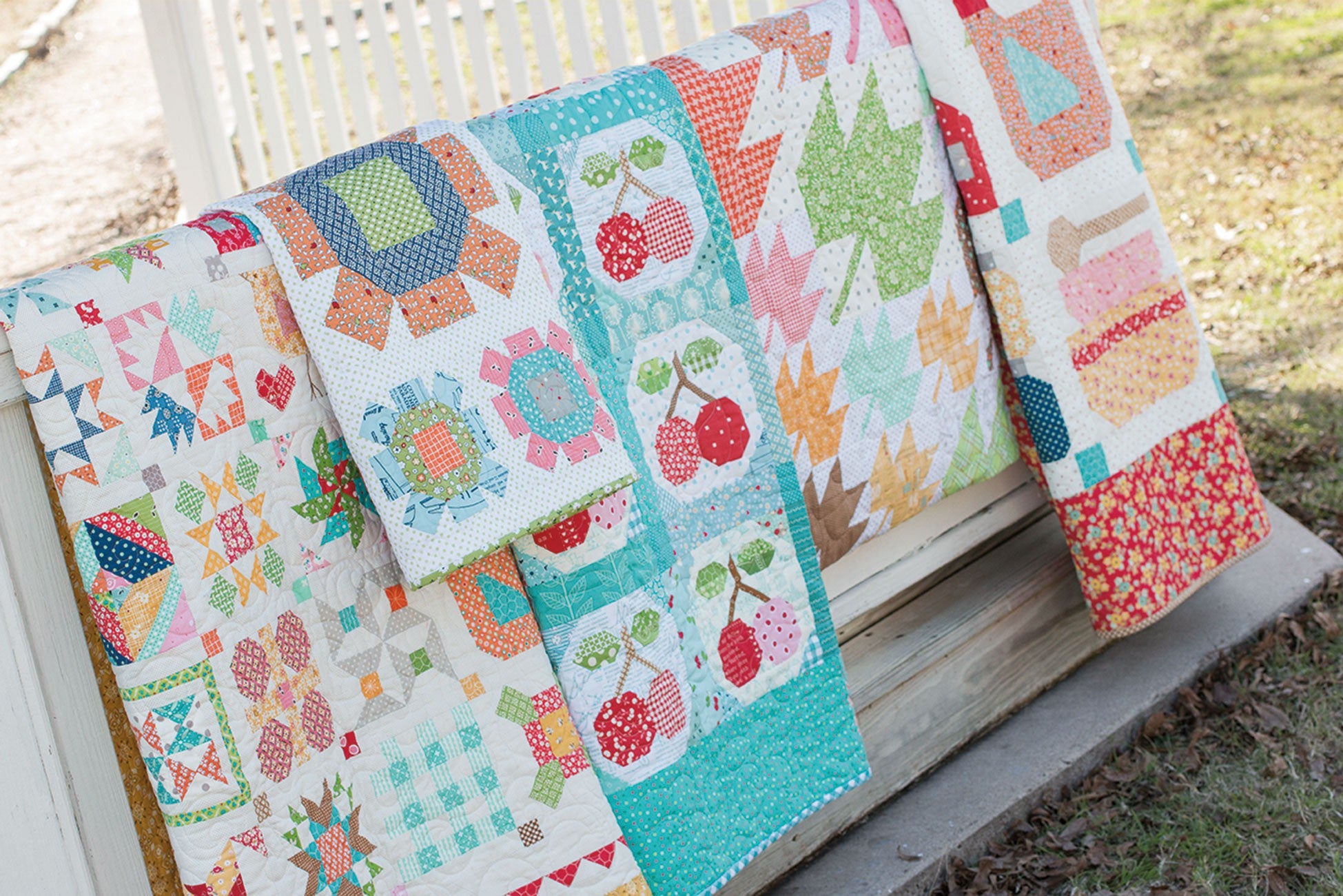 Several Farm Girl Vintage quilts by Lori Holt for It's Sew Emma draped over a railing by a white picket fence, including a sampler, a cherries quilt, and a scrappy maple leaf quilt.