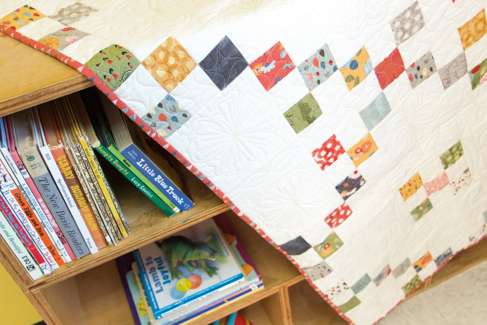 Interior quilt from Precut Primer, scattered charm squares on a white background beside a bookshelf.