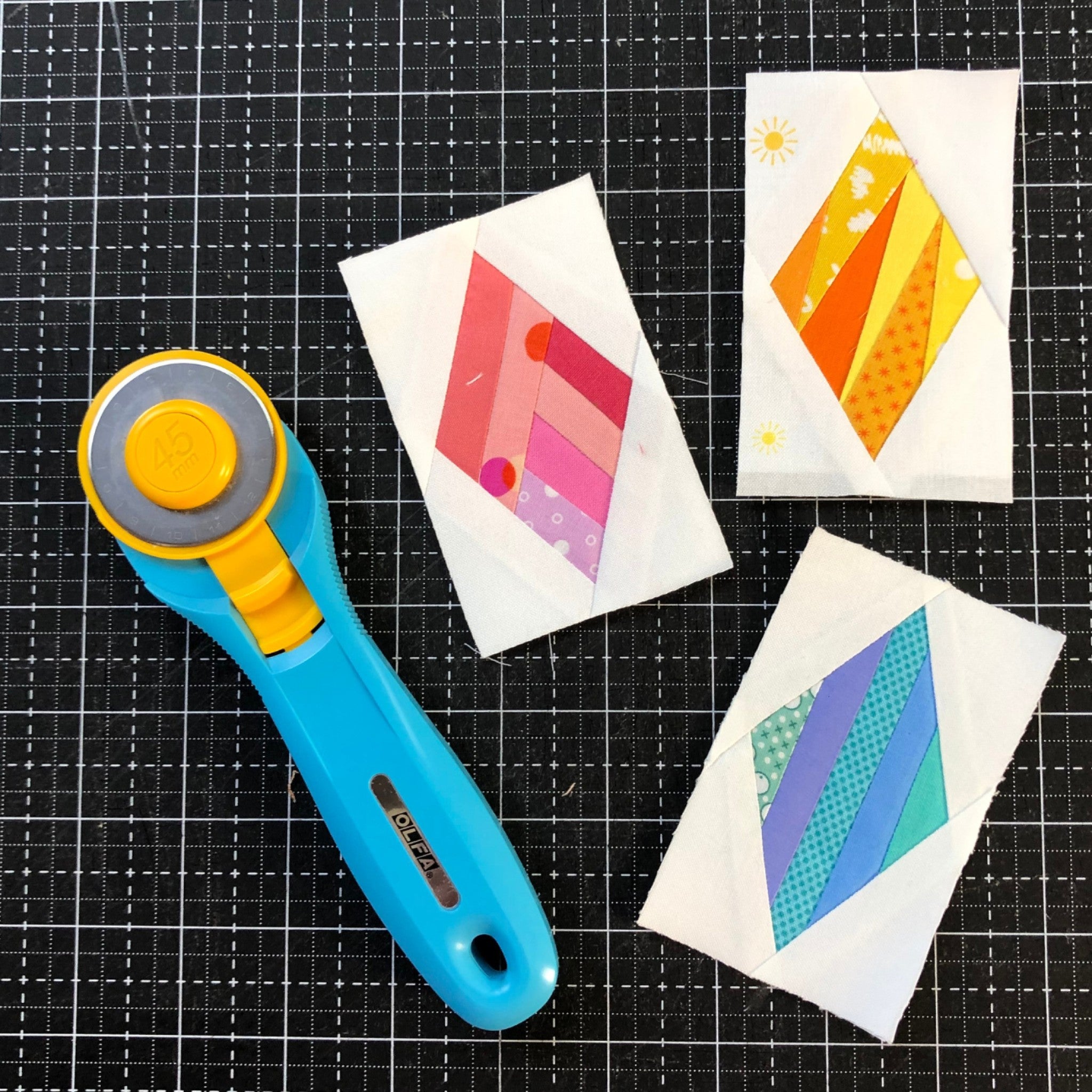 Three finished Mini Diamond blocks from Leila Gardunia's foundation paper pad, arranged on a black cutting mat beside a blue and yellow rotary cutter. One diamond is pieced in pink and red stripes, one in orange and yellow prints including a sun motif, and one in blue, green, and teal. Each diamond is set on a white background, showing the 2 inch by 3 1/2 inch finished block size.