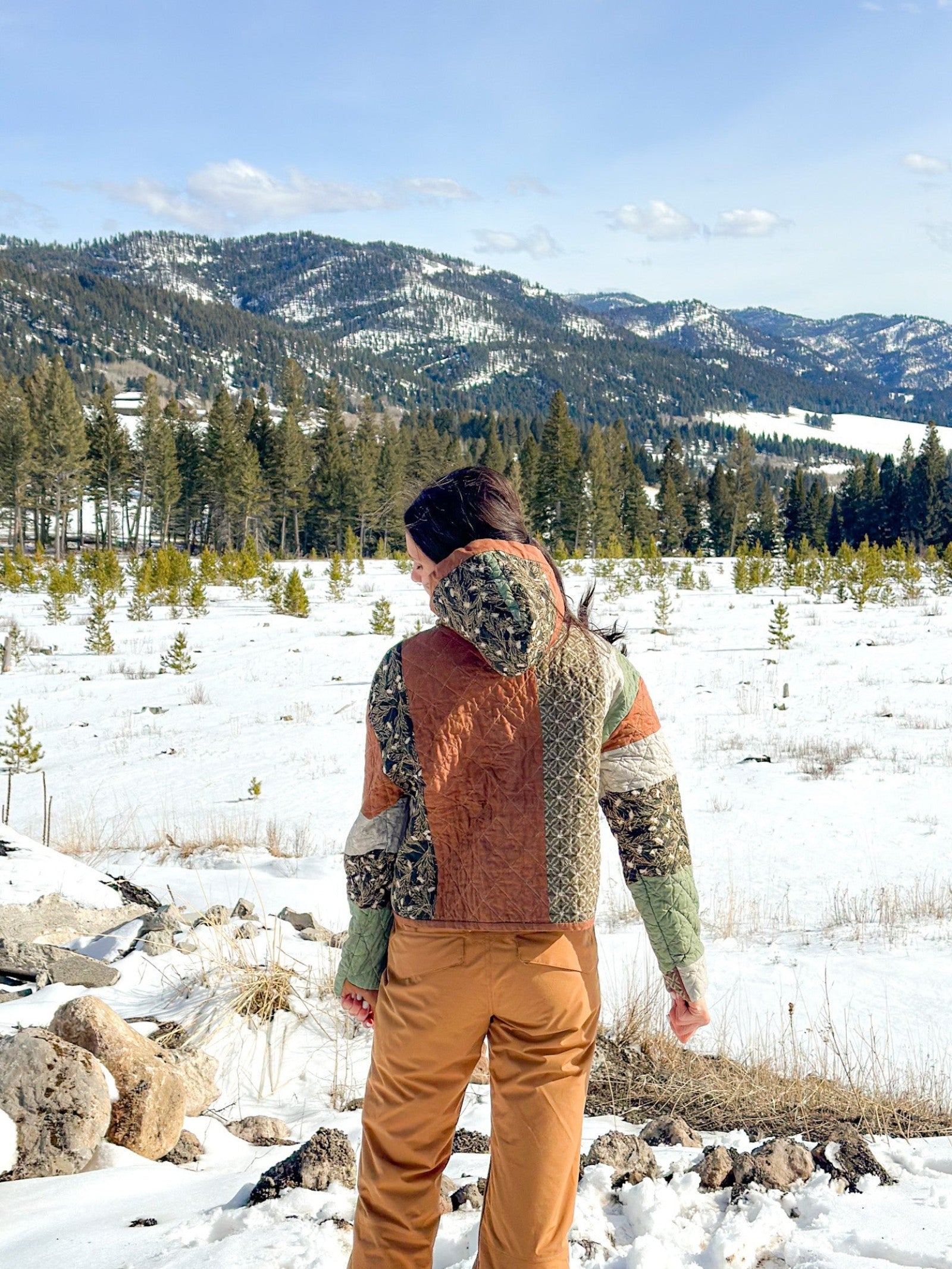Back view of a person wearing the finished Quilted Hoodie in a green, rust, and cream patchwork colorway, showing the quilted back panels and lined hood resting down, standing in a snowy mountain landscape.
