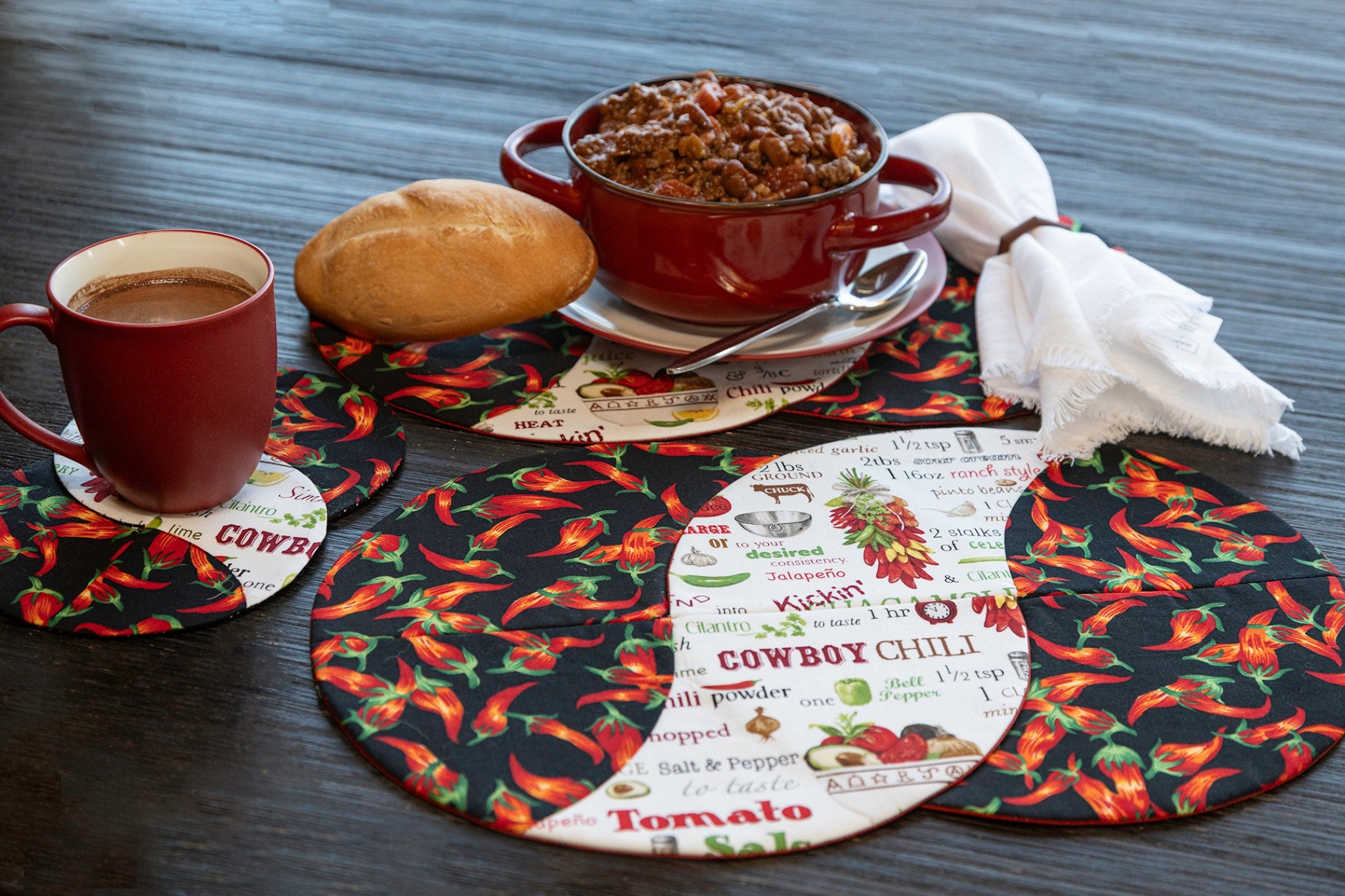 Braided Twist Junior placemats and Half Pint mug rug in red chili pepper print and Cowboy Chili recipe novelty fabric, styled on a dark wood table with a red ceramic bowl of chili, a crusty bread roll, white napkin, and a red coffee mug.