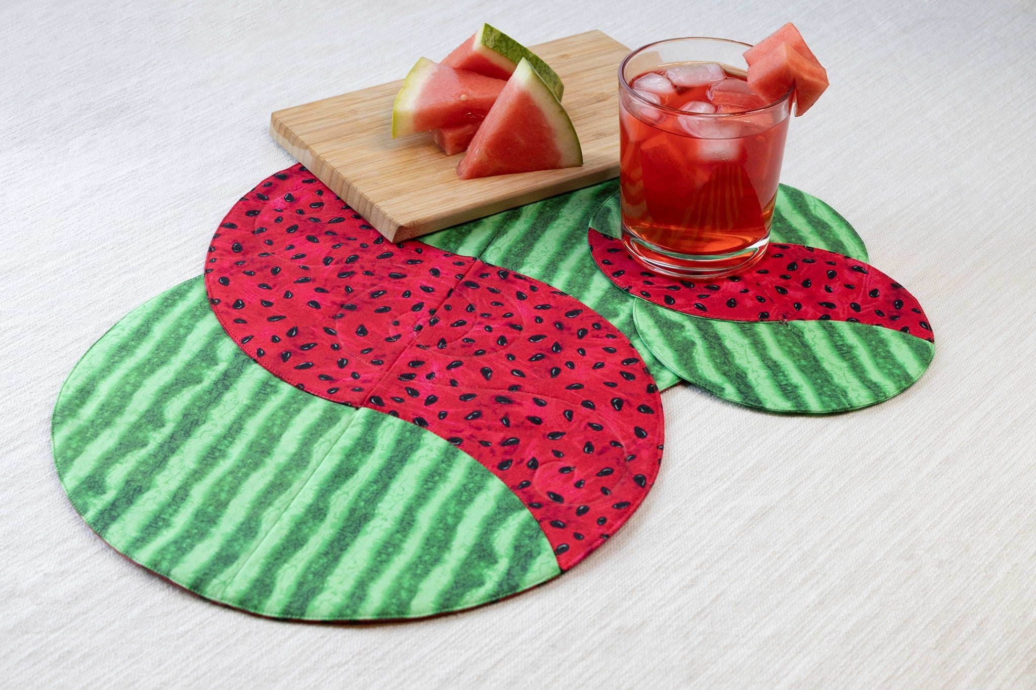 Braided Twist Junior placemat and Half Pint mug rug in watermelon-themed quilting cotton, with bold red watermelon flesh prints featuring black seeds in the curved center sections and green rind with darker stripes on the outer crescents. Styled on a cream linen surface with a bamboo cutting board holding fresh watermelon slices and a clear rocks glass of iced watermelon drink garnished with watermelon chunks.