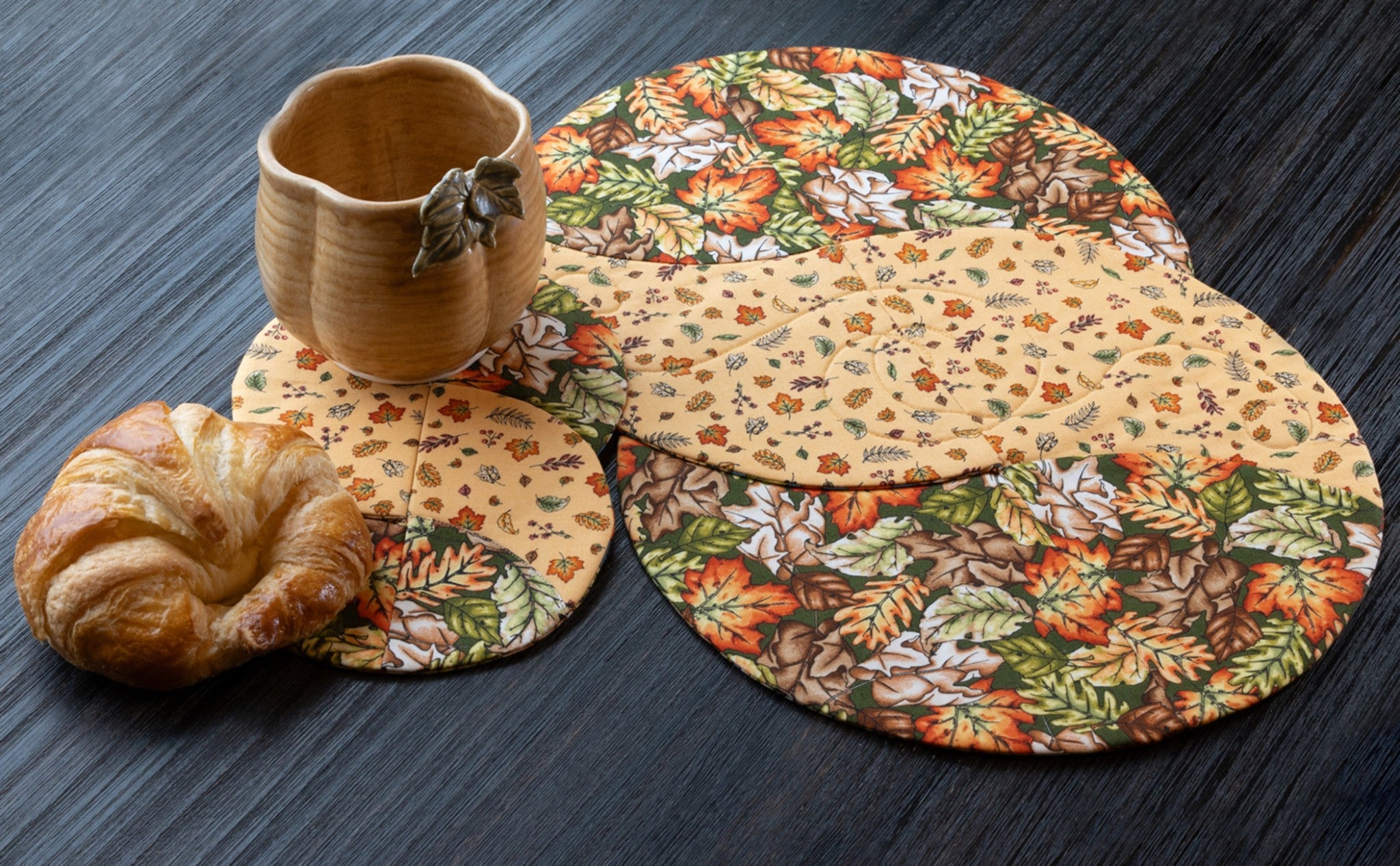 Braided Twist Junior placemats and Half Pint mug rug in autumn prints, styled on a dark wood table. A pumpkin-shaped buttery cream ceramic mug topped with a small green leaf accent sits beside a flaky croissant. The placemat and mug rug feature bold large oak and maple leaves in orange, gold, green, and brown on a dark green ground and a small-scale autumn leaf and acorn print on a soft butter yellow background.