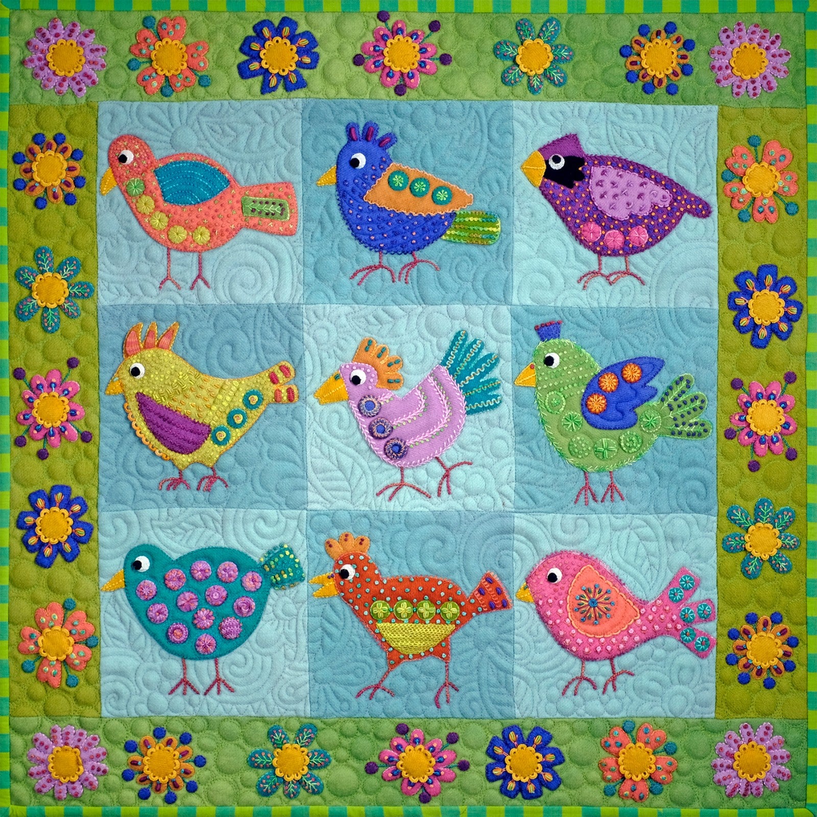 The finished Chirp wall hanging by Sue Spargo of Folk Art Quilts, filling the frame. Nine wool birds in coral, blue, purple, yellow, green, pink and teal stand in a grid on quilted light blue blocks, surrounded by a green border of layered wool flowers with embroidered detail.