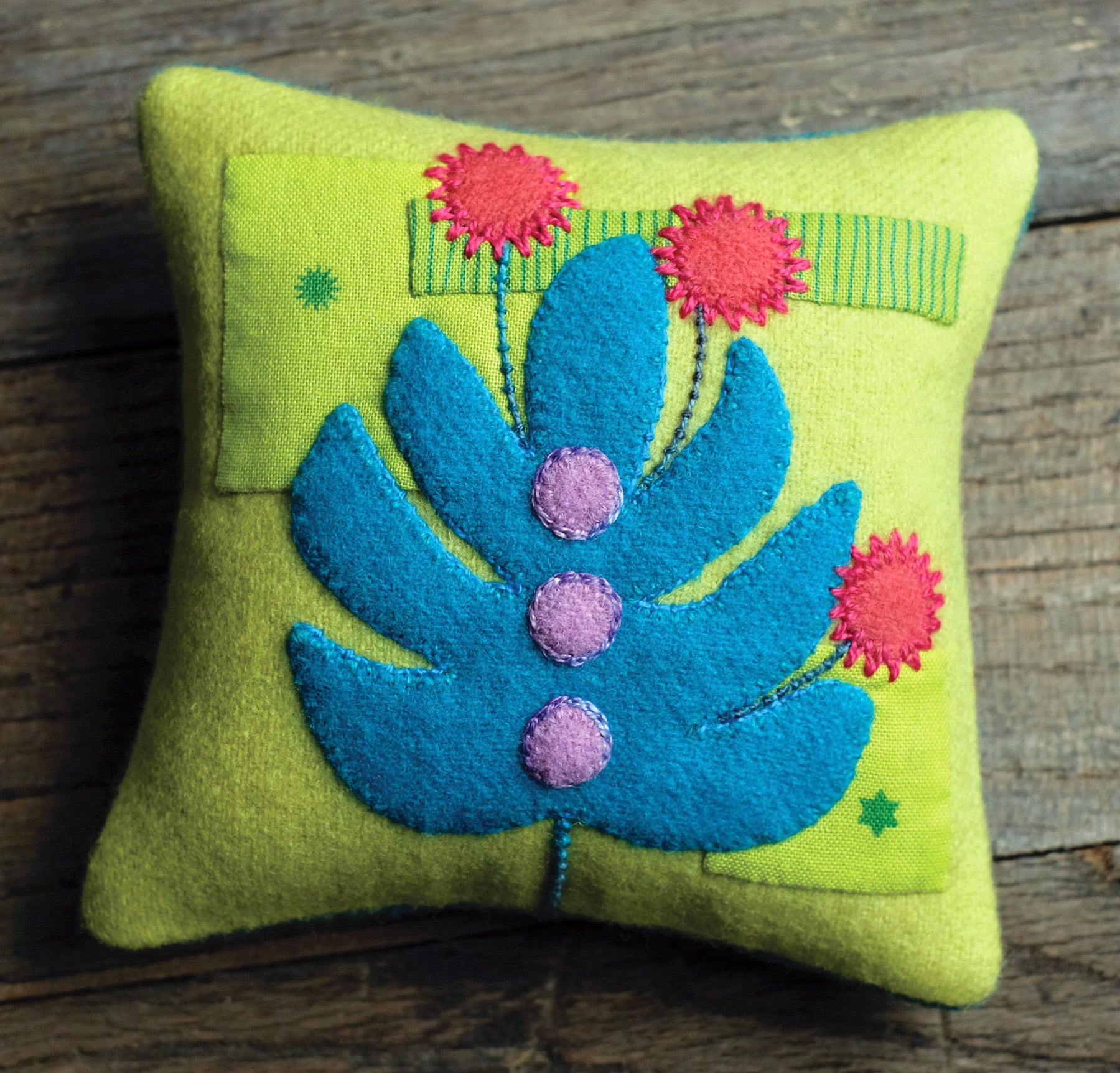 The Breadfruit Pincushion, one of the book's bonus projects, a green wool pincushion with a blue breadfruit leaf and red flowers in wool applique and embroidery.