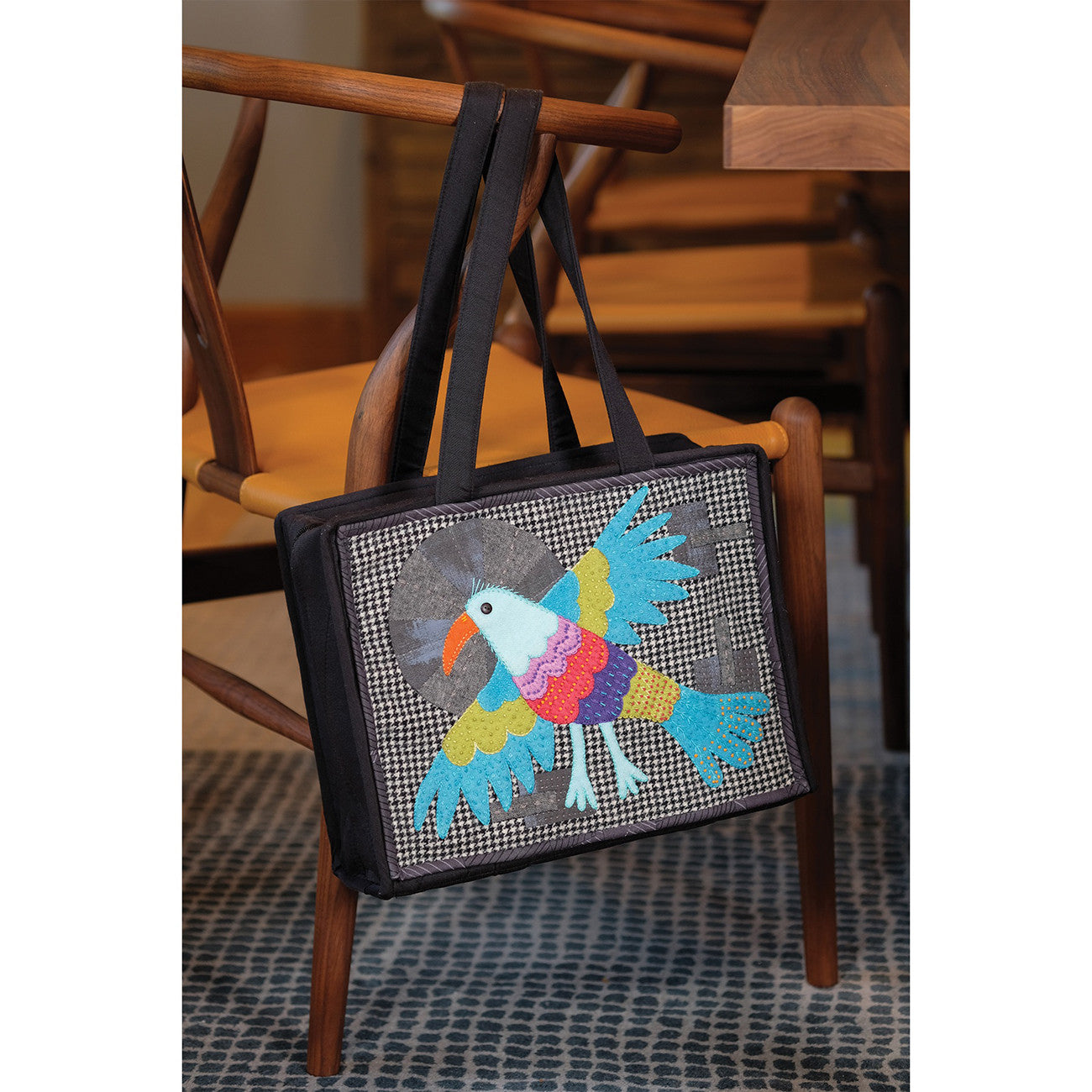 The Of a Feather Workbox, a bonus project, a fabric project bag with a bright folk art bird appliqued on the front, shown hanging on a chair.
