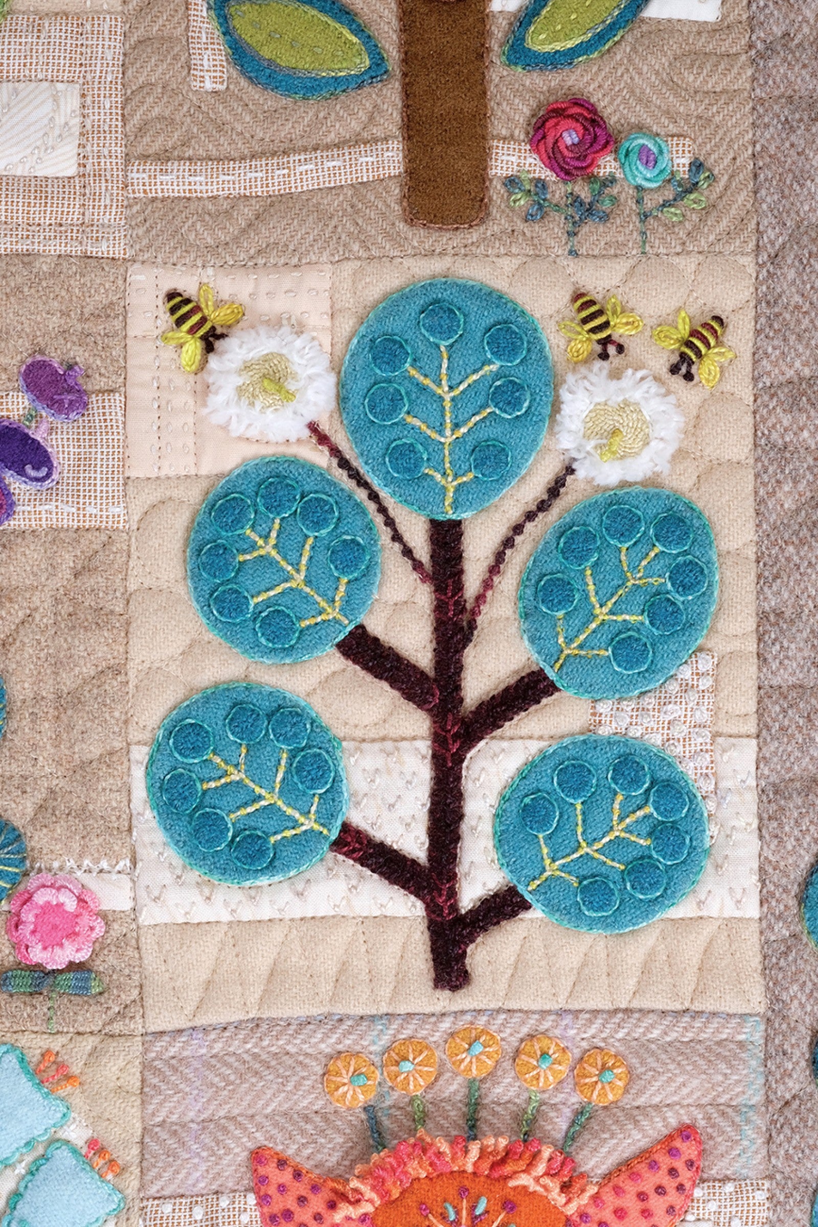 Close detail of a Rooted block by Sue Spargo of Folk Art Quilts. A tree with round teal wool leaves and a dark trunk is surrounded by stitched bees and orange flowers, in layered applique and dense hand embroidery on a quilted background.