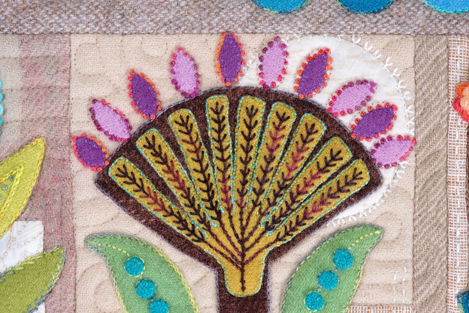 Close detail of a Rooted block by Sue Spargo of Folk Art Quilts. A fan-shaped tree in gold and olive wool with purple petal tips and teal accents, worked in layered applique and hand embroidery on a quilted neutral background.