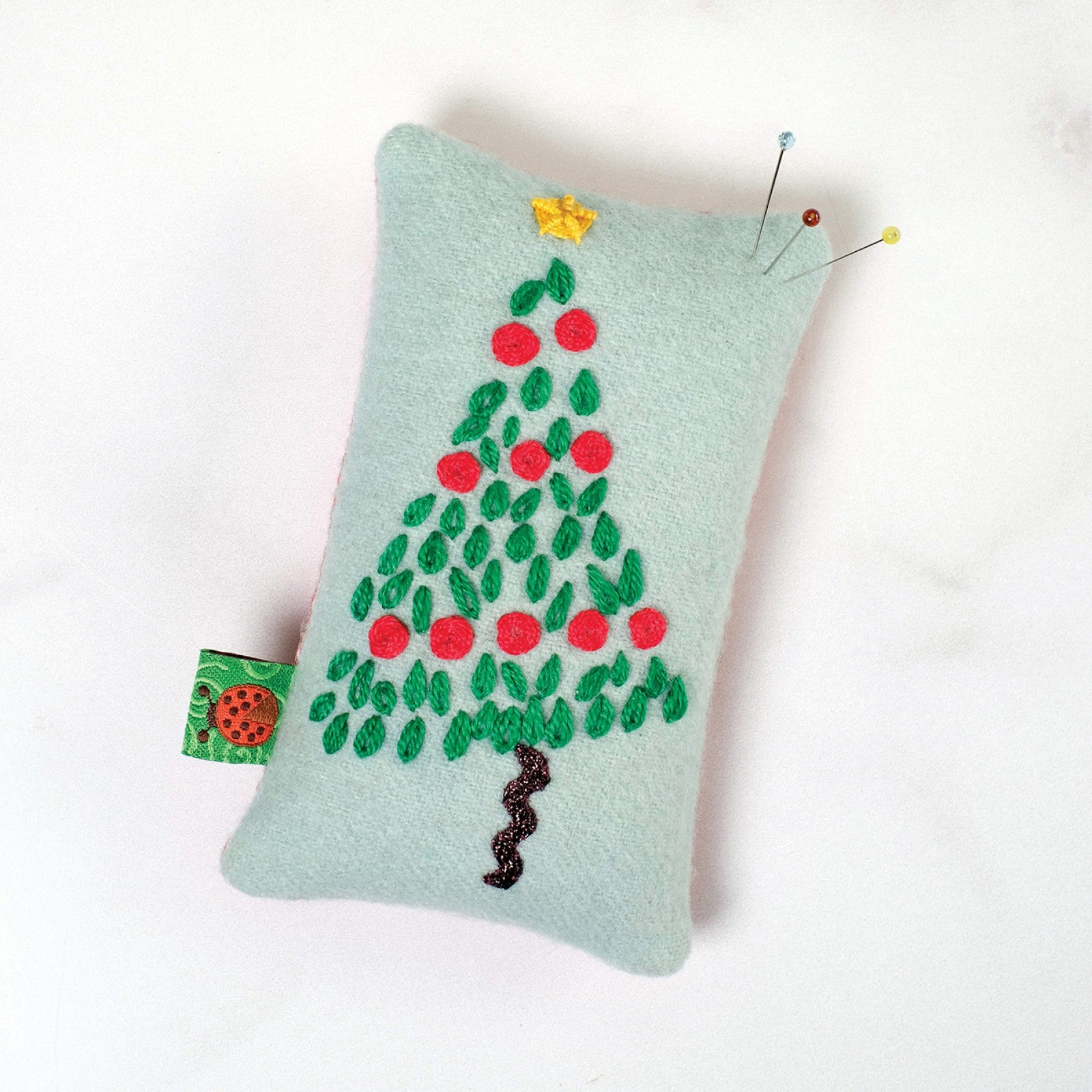 The bonus Christmas Tree pincushion from Tinsel by Sue Spargo of Folk Art Quilts, a pale green wool pincushion with an embroidered and beaded Christmas tree and a small fabric tab at one corner.