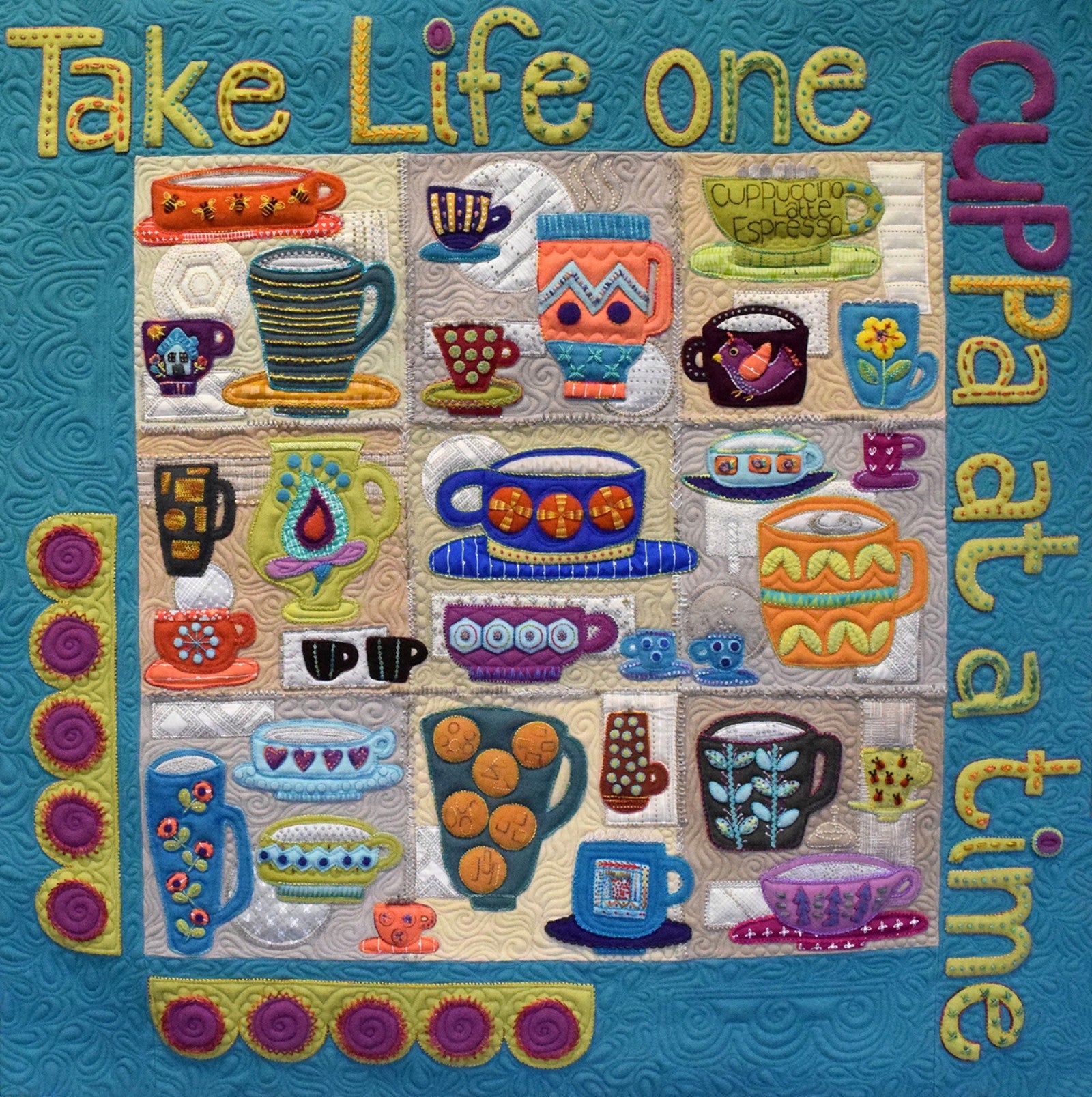 The finished Cuppa quilt by Sue Spargo, a grid of wool applique teacups and mugs with embroidery and button details on a teal ground, bordered by the words Take Life one cuppa at a time.