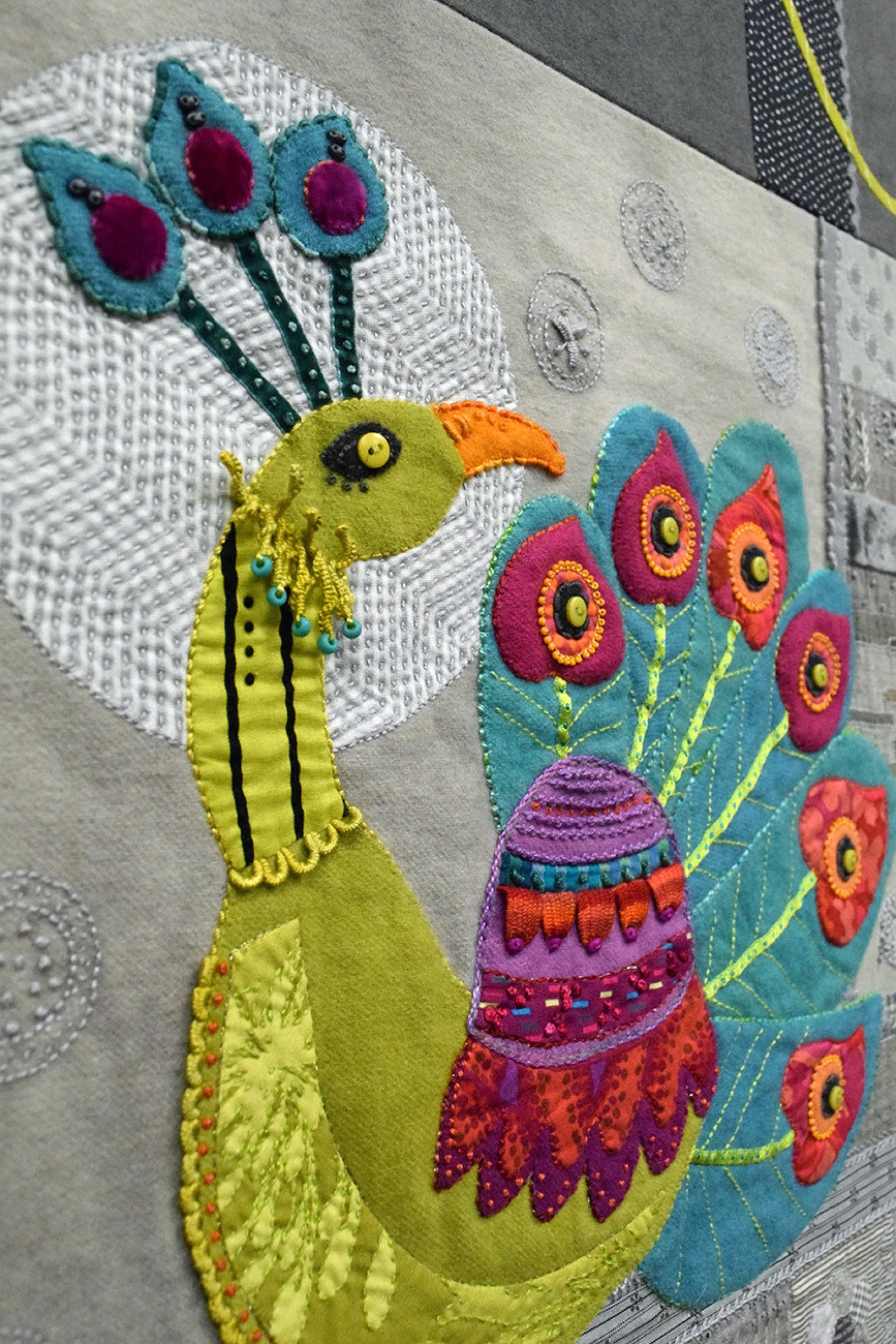 Peacock Block quilting pattern by Sue Spargo of Folk Art Quilts. Close-up of the appliqued peacock showing the beaded topknot, button eye, layered wool and velvet body and tail feathers, and the seed stitching and French knots filling the background.