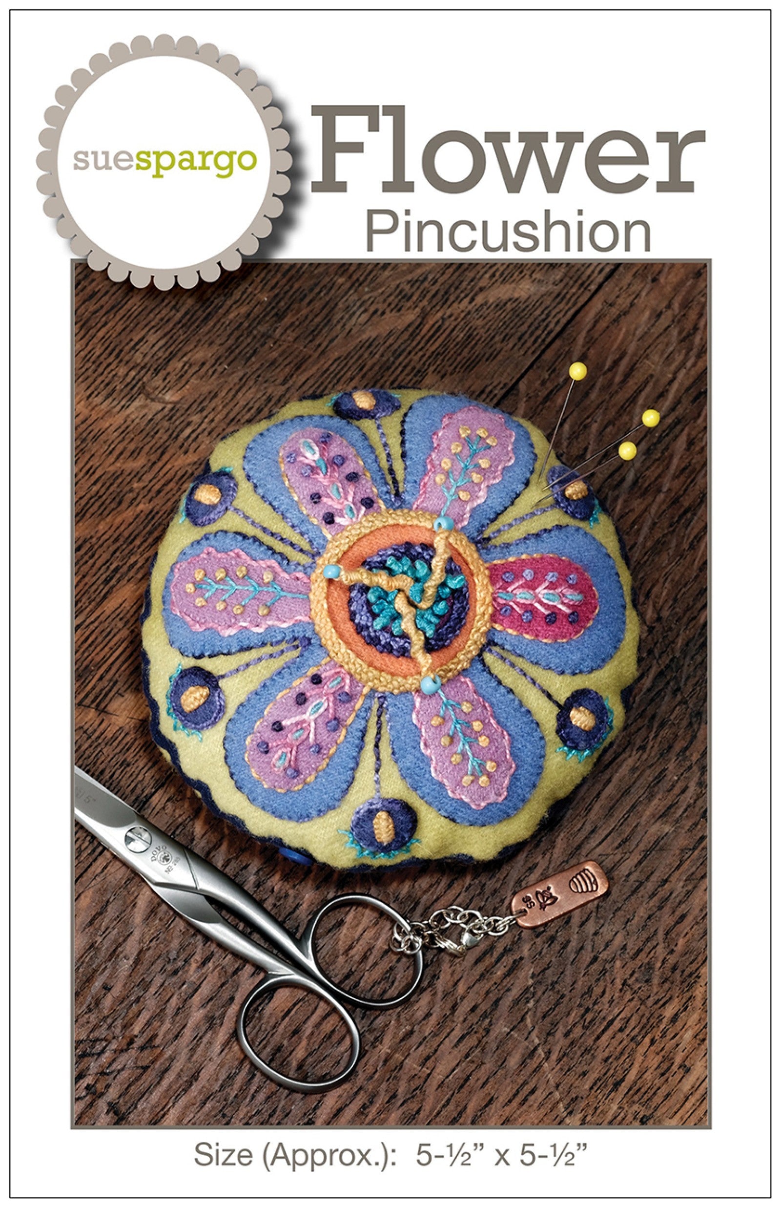 Flower Pincushion sewing pattern by Sue Spargo of Folk Art Quilts. Cover shows the finished round pincushion: a large folk-art flower with layered periwinkle and lavender petals and a coral center on a green wool ground, worked in wool appliqué with dimensional embroidery and a scalloped edge. Size about 5-1/2 inches across shown.