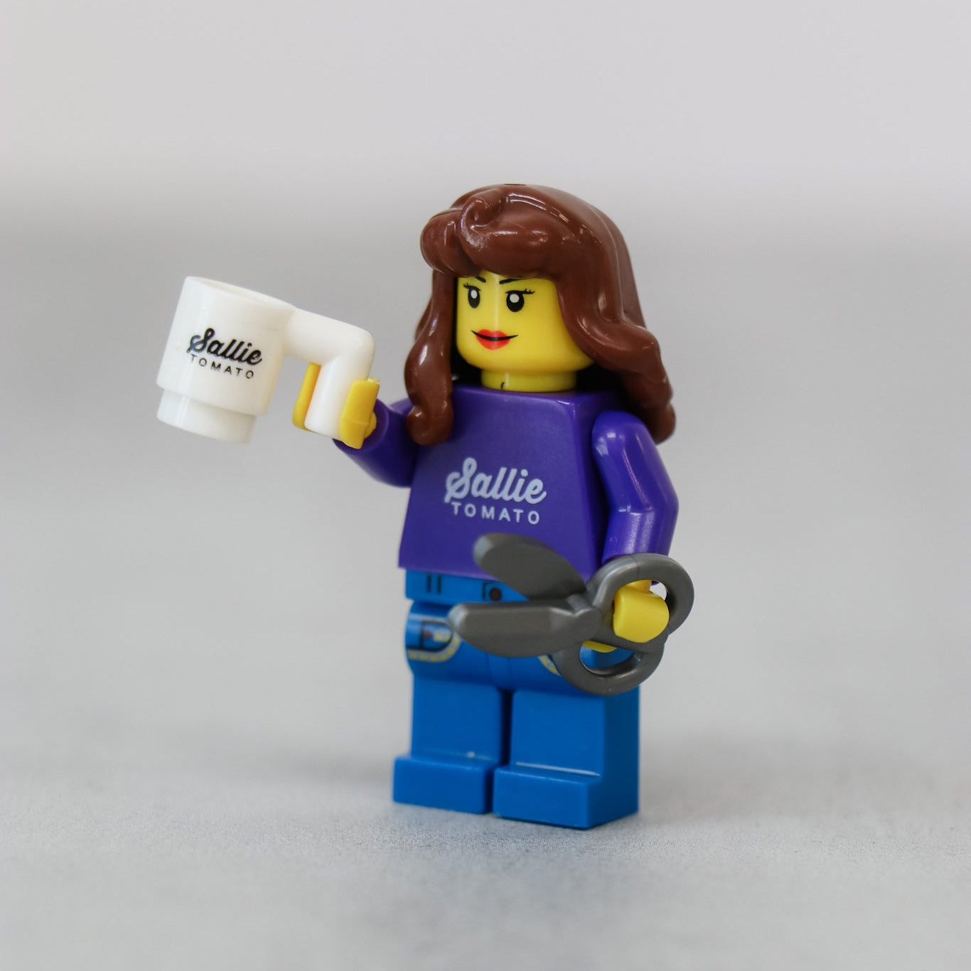 Close-up of the sewist figurine from the Sallie Tomato Sewist Building Block Set, with brown hair and a purple Sallie Tomato shirt, holding an oversized pair of scissors.