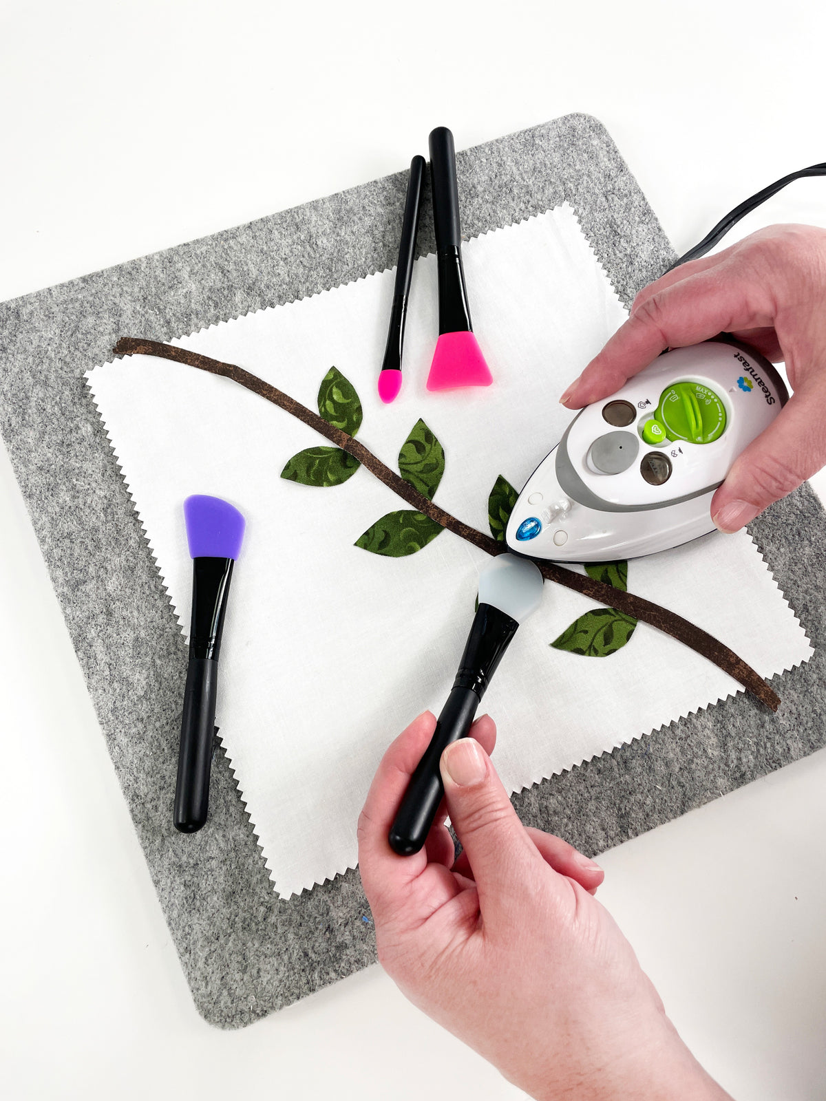 Overhead view of Position and Press Silicone Tools in use on a gray wool pressing mat: one hand holds appliqué in place with a silicone tip while the other guides a mini iron.