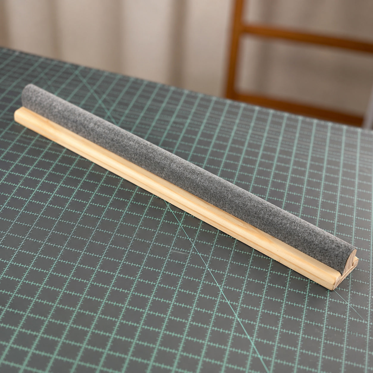 The Gypsy Quilter 20-inch Wool Pressing Bar laid diagonally on a green cutting mat in a sewing room, the long gray wool-covered rounded top mounted along a light wood clapper base.
