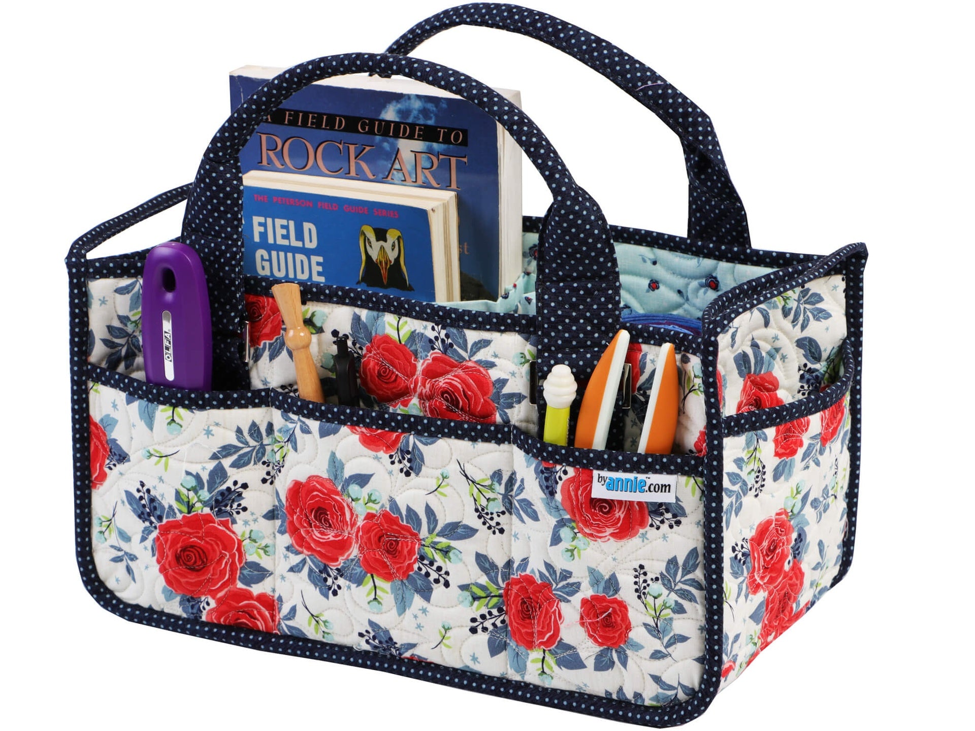 Catch All Caddy 2.0 sewn in a navy and red rose print, standing upright and filled with field guides, a marker, and craft tools tucked into the exterior slip pockets, with padded fabric handles.