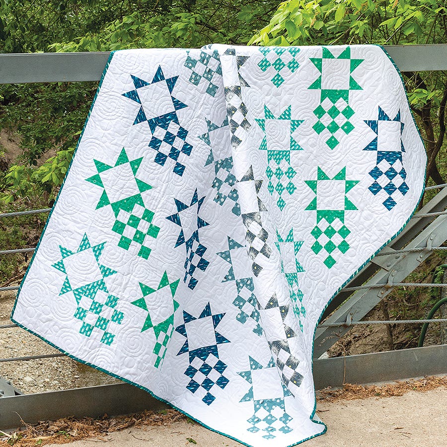 Comet Quilt from Simply Half Yards, teal and aqua star blocks on a white background, draped on a fence.